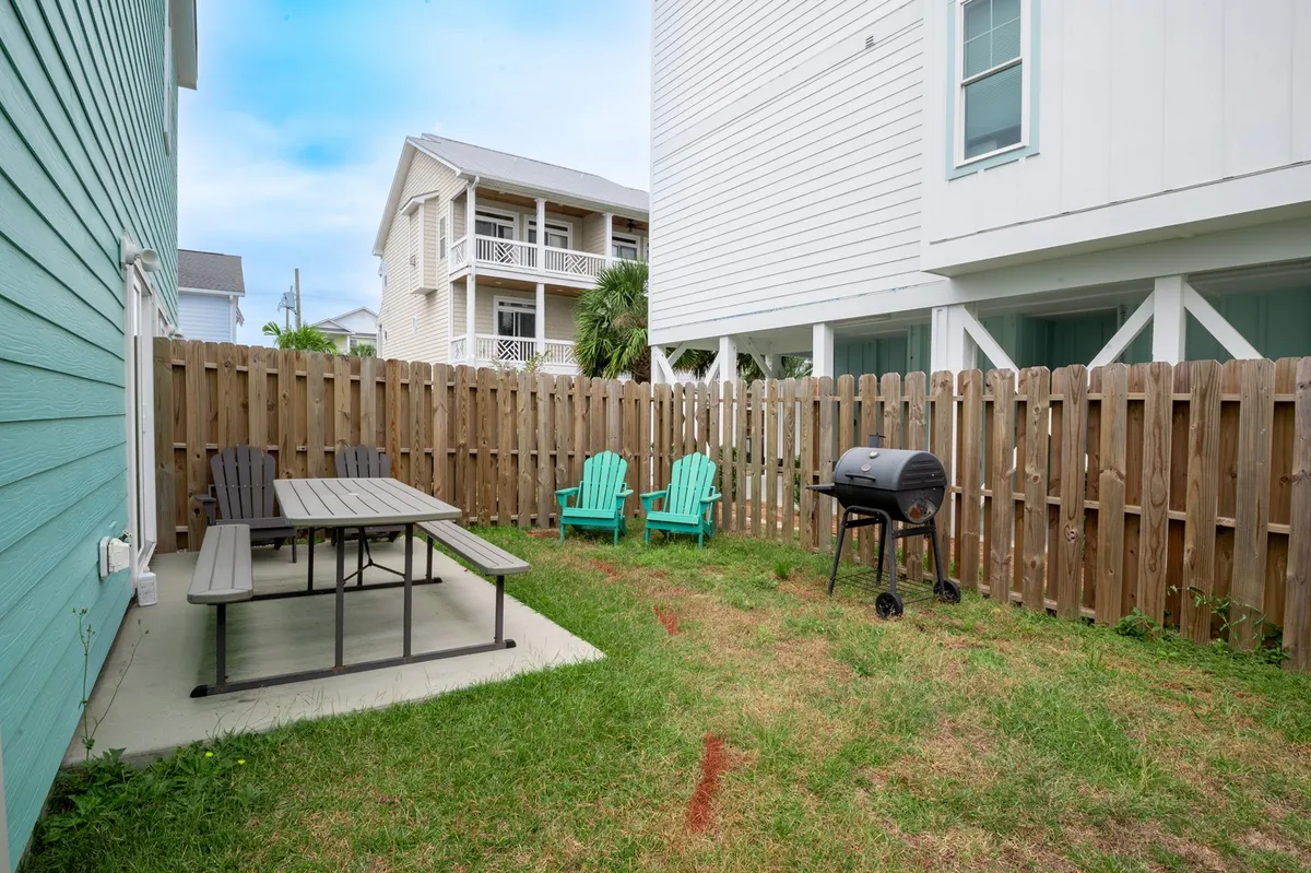A spacious fenced backyard featuring a wooden picnic table, a propane grill, adirondack chairs, and a tall privacy fence. The setting is casual and perfect for group gatherings, with plenty of open grassy space for kids and pets to play safely.