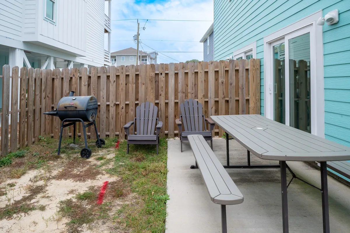 An inviting backyard scene featuring a grill, Adirondack chairs, and a picnic table set up for outdoor dining. The space is designed for group gatherings, relaxation, and family barbecues, with the beach atmosphere subtly present.