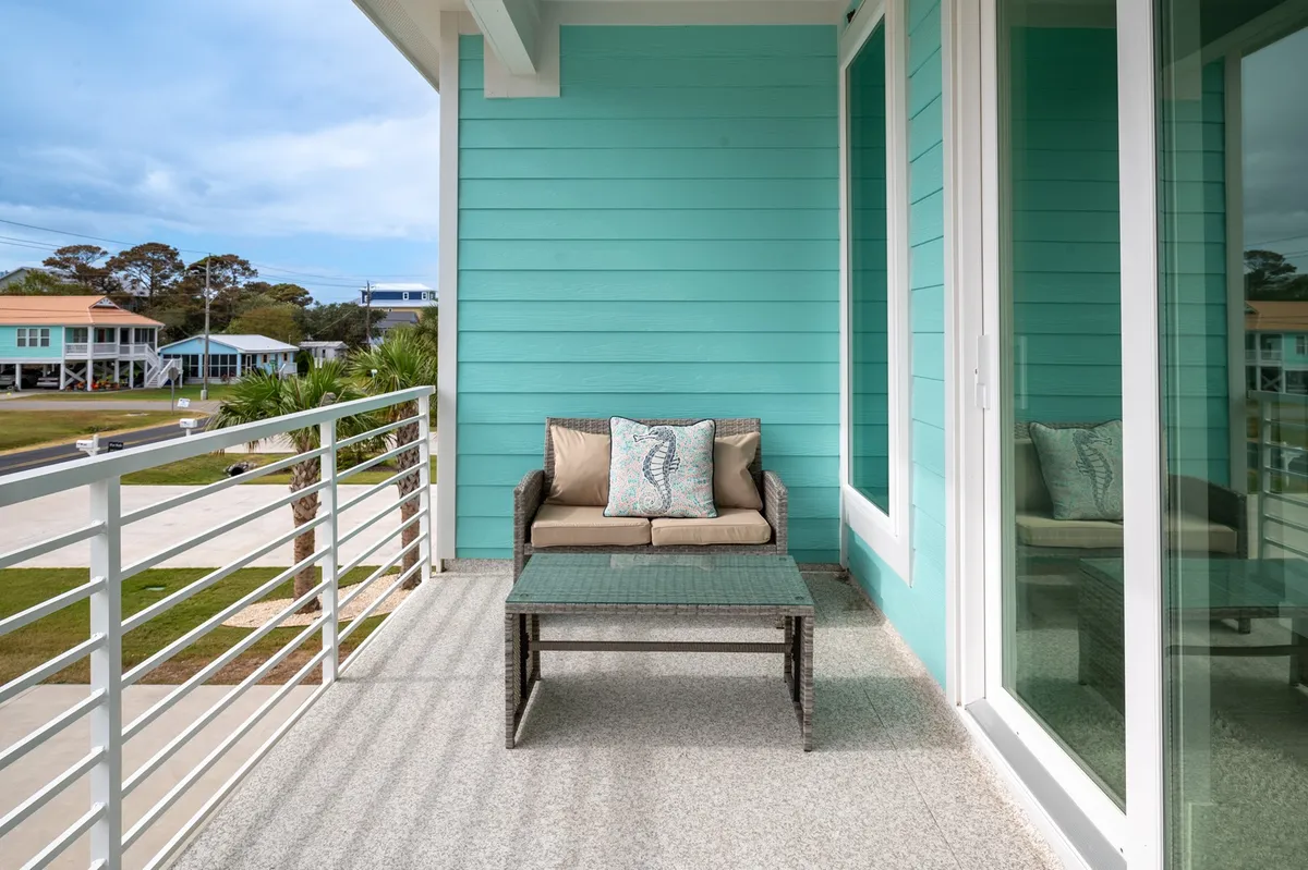The image depicts an inviting outdoor seating area against a cheerful turquoise wall. Cushioned chairs and a decorative pillow create a relaxing spot for enjoying the outdoors. The setting suggests tranquil moments at sunrise or in the evening, evoking the coastal lifestyle and the ease of transitioning between indoor comfort and outdoor pleasure.