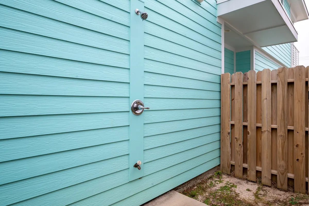 This image features a vibrant turquoise wall, a sturdy wooden fence, and an outdoor metal door handle. The scene is centered around the outdoor shower area, offering a sense of privacy and fun. The lively colors and private setup are perfect for rinsing off a sandy dog after the beach.