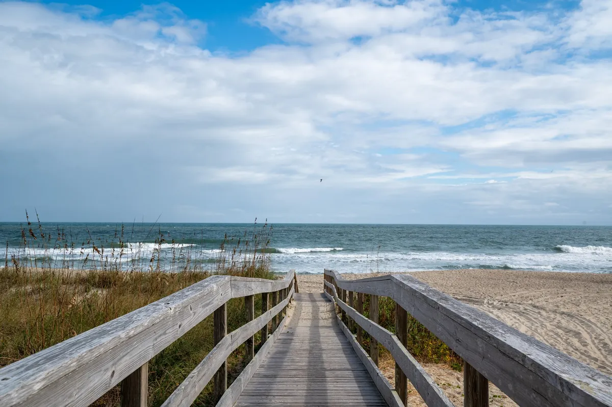 A scenic exterior shot of a wooden boardwalk stretching towards the ocean, with rolling waves, dune grasses, and a partly cloudy sky. The tranquil setting illustrates the nearby beach environment, inviting guests (and their furry friends) to explore and enjoy the coast just a stroll away from Tide and Seek.