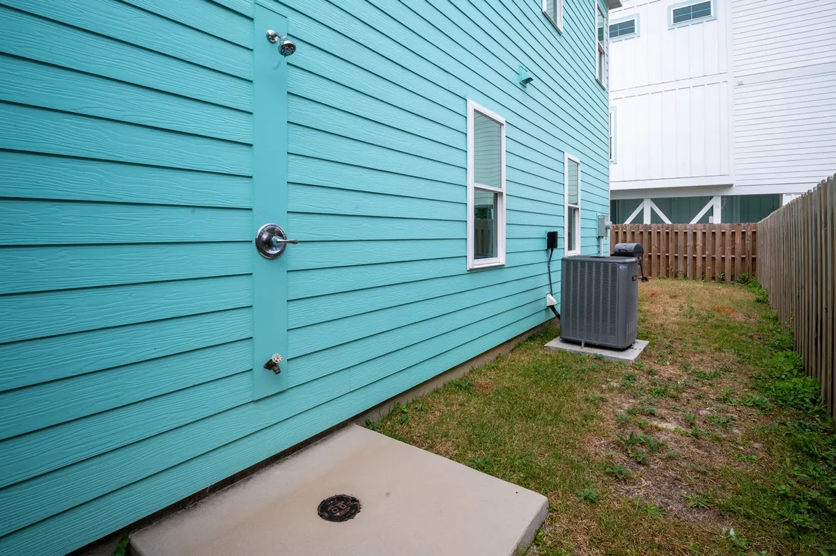 A vibrant turquoise exterior wall with an outdoor shower, window, and nearby yard space. The high wooden fence offers privacy, and the shower is conveniently located for rinsing off after a day at the beach.