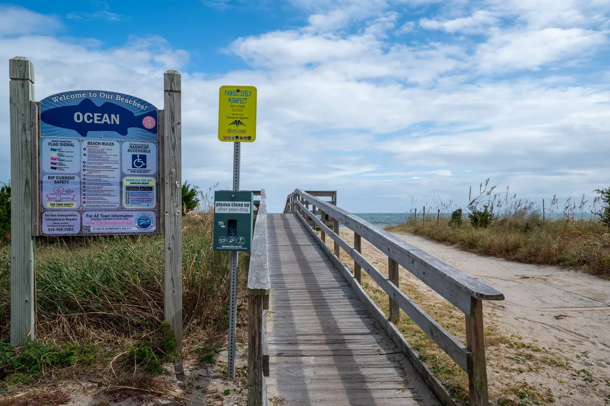 This image shows a welcoming boardwalk leading directly to the sandy beach, with a clear ocean view in the background. A 'welcome' sign greets visitors, and the relaxed atmosphere is accentuated by soft light and open skies.