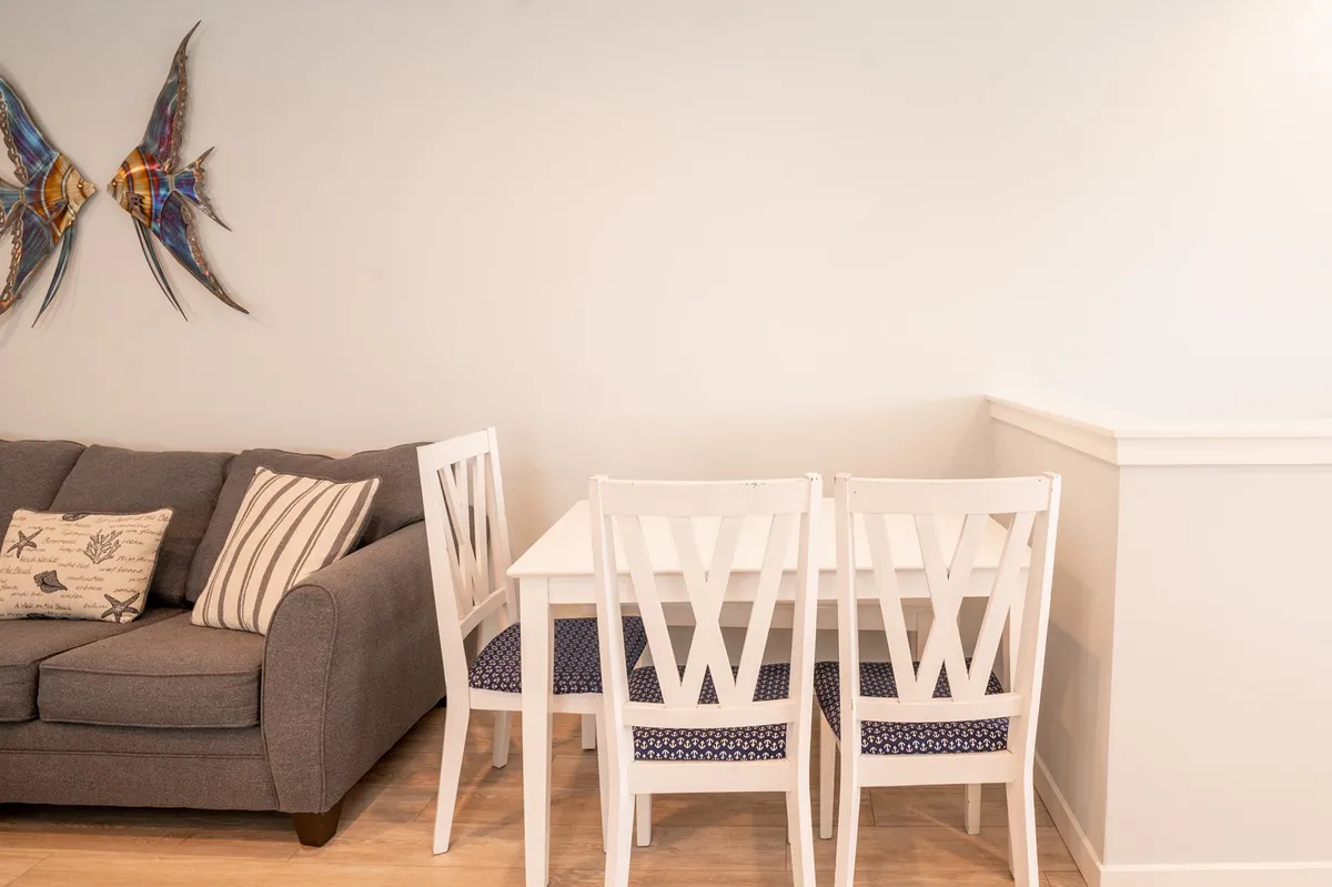 A cozy dining area with a white dining table, surrounded by comfortable seating and decorative coastal artwork. The space is bathed in natural light and is designed for shared meals, casual gatherings, and relaxed celebrations with friends and family.