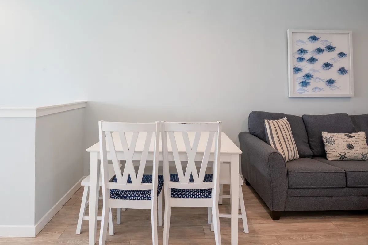 A fresh, open dining area with a white dining table, blue upholstered chairs, a gray sofa in the background, and framed artwork on the wall. The space is bright and welcoming, designed for shared meals, games, or casual conversations.