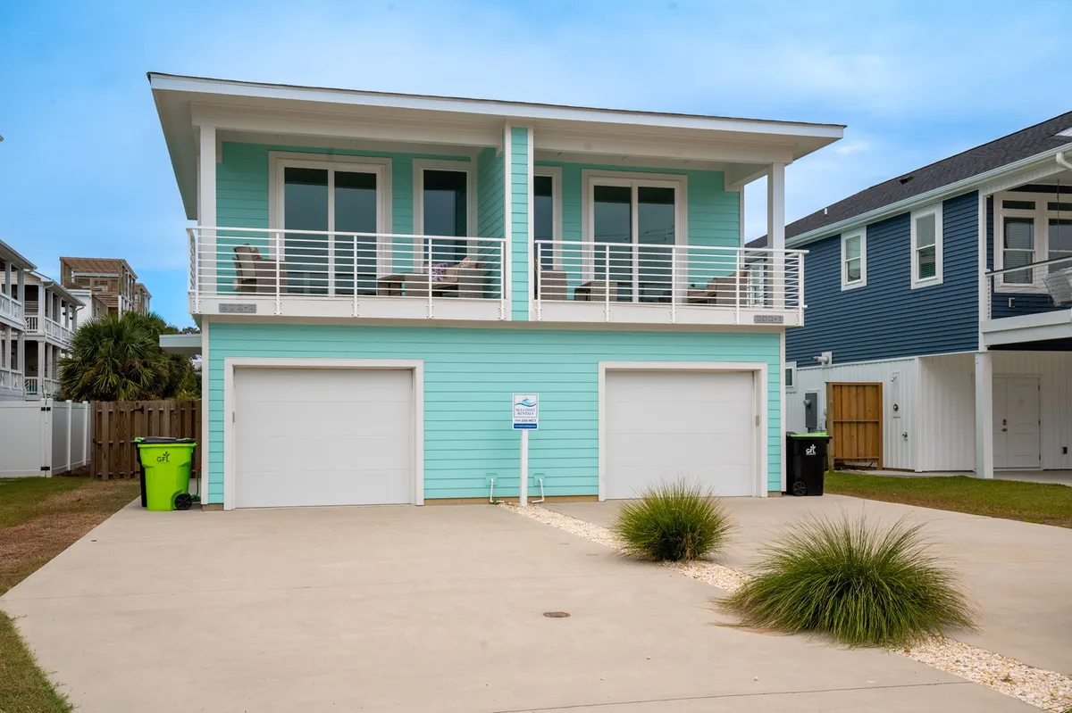 The image showcases a cheerful turquoise beachfront condo with spacious balconies furnished with inviting chairs, double garage doors, and a sunlit yard perfect for outdoor activities. The scene exudes a welcoming vibe, hinting at sunset balcony dinners, morning coffee with ocean views, and room for relaxation and play.
