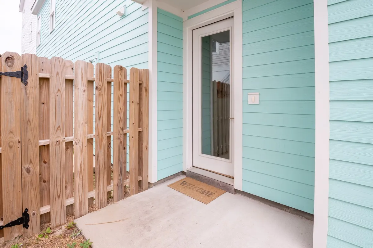 The image shows the welcoming front entrance of Tide and Seek, with blue exterior walls, a sturdy wooden fence, and a front door featuring a glass panel. The welcome mat and casual outdoor setting create an inviting first impression, and the visible fence hints at the secure, private yard ideal for dogs to play safely.