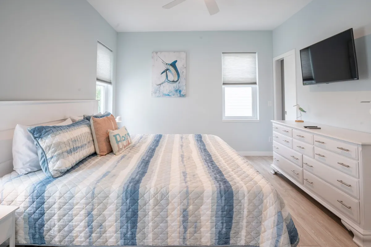 A tranquil bedroom with a bed dressed in a striped quilt, whimsical fish-themed wall art, a white dresser, and a wall-mounted television. The atmosphere is peaceful and cozy—ideal for relaxing, reading, or enjoying a quiet evening.