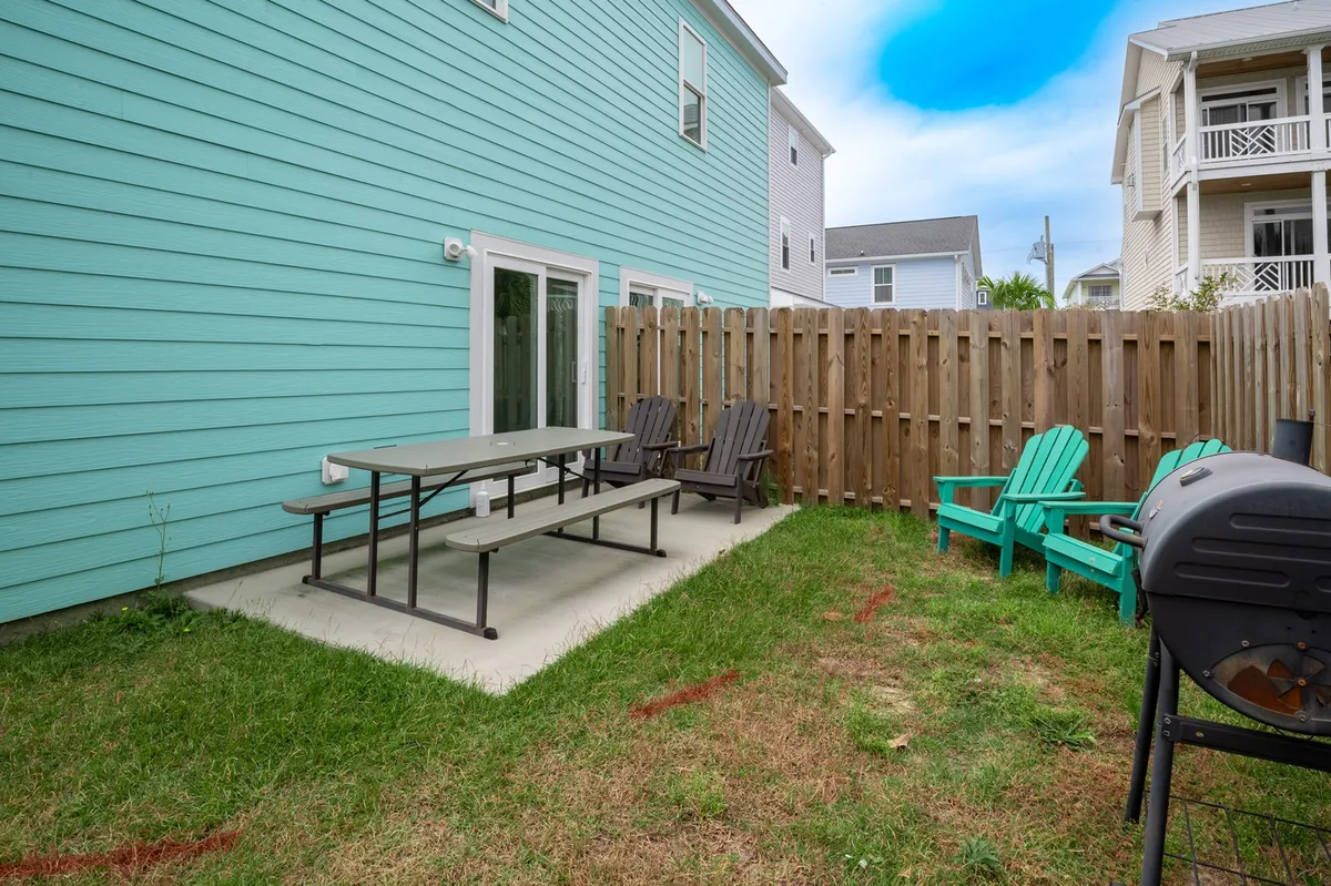 Another angle of the fenced backyard showing a picnic table, adirondack chairs, and a propane grill. The space is bathed in natural light and invites relaxation and social gatherings, highlighting the ease of outdoor entertaining.